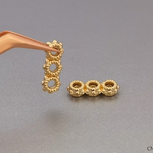 Puede incluir: Separadores de cuentas de metal dorado con un diseño circular texturizado. Los separadores se muestran sostenidos por unas pinzas.