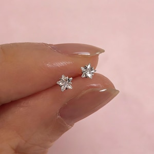 May include: A pair of small, silver-colored stud earrings. Each earring features a flower design with multiple clear, sparkling stones. The earrings are displayed against a soft pink background.