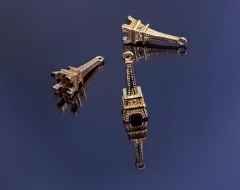 May include: Three gold-colored Eiffel Tower charms are displayed against a dark blue background. The charms are detailed with intricate designs and have a loop at the top for attaching to jewelry. The charms are reflective.