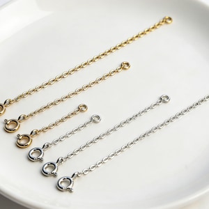 May include: An assortment of gold and silver-colored chain extenders with spring ring clasps. The extenders are of varying lengths and are arranged on a white, slightly textured surface. The chains are made of small, connected circular links.