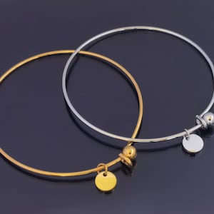 May include: Two thin, minimalist bangles. One is gold-toned, the other is silver-toned. Each has a small, round charm and a decorative ball. The bangles are displayed on a dark, reflective surface. Jewelry.