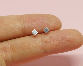 May include: A pair of small, square-shaped silver stud earrings. The earrings are displayed on a pink background, held by a person's fingers. The earrings have a simple, minimalist design.
