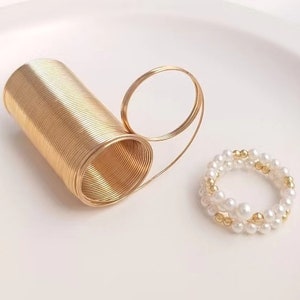 May include: Two gold rings, one is a simple band and the other is a coiled wire ring. The coiled wire ring is open and can be adjusted to fit different finger sizes. The other ring is made of white pearls and gold beads.