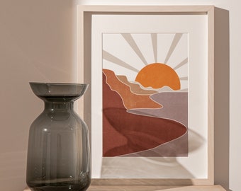 May include: A dark gray glass vase with a wide mouth sits on a light wood table in front of a framed print of a sunset landscape with orange, brown, and beige colors.