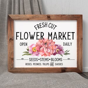 May include: A white sign with a wooden frame, featuring the text "FRESH CUT FLOWER MARKET OPEN DAILY" and a floral design with pink and purple flowers. The sign also includes the text "SEEDS STEMS BLOOMS ROSES. PEONIES. TULIPS AND DAISIES."