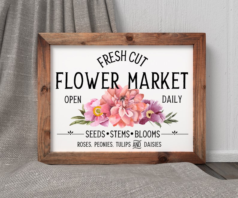 May include: A white sign with a wooden frame, featuring the text "FRESH CUT FLOWER MARKET OPEN DAILY" and a floral design with pink and purple flowers. The sign also includes the text "SEEDS STEMS BLOOMS ROSES. PEONIES. TULIPS AND DAISIES."