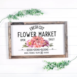 May include: A rustic wooden sign with the text "FRESH CUT FLOWER MARKET" in black lettering. The sign features a watercolor illustration of pink and purple flowers with green leaves. The text "OPEN" and "DAILY" are also included, along with the phrase "SEEDS STEMS BLOOMS ROSES, PEONIES, TULIPS AND DAISIES."