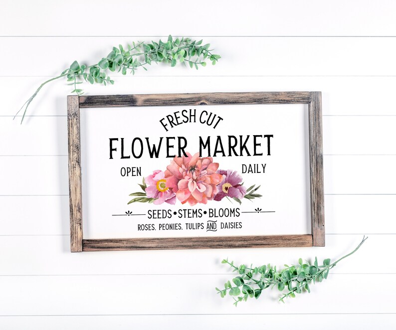 May include: A rustic wooden sign with the text "FRESH CUT FLOWER MARKET" in black lettering. The sign features a watercolor illustration of pink and purple flowers with green leaves. The text "OPEN" and "DAILY" are also included, along with the phrase "SEEDS STEMS BLOOMS ROSES, PEONIES, TULIPS AND DAISIES."