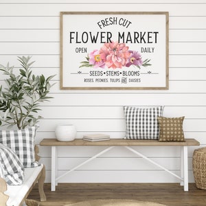 May include: A white sign with a wooden frame reads "FRESH CUT FLOWER MARKET" with pink and purple flowers in the center. The sign also says "OPEN DAILY" and "SEEDS STEMS BLOOMS ROSES. PEONIES. TULIPS AND DAISIES".