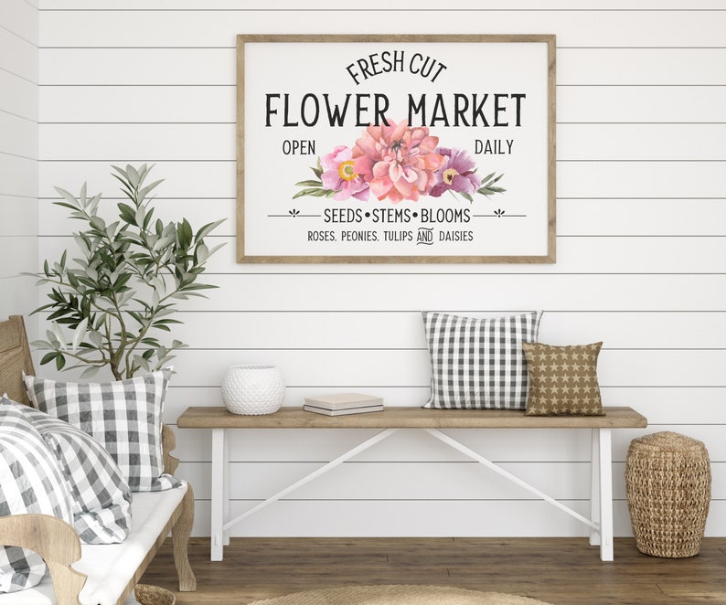 May include: A white sign with a wooden frame reads "FRESH CUT FLOWER MARKET" with pink and purple flowers in the center. The sign also says "OPEN DAILY" and "SEEDS STEMS BLOOMS ROSES. PEONIES. TULIPS AND DAISIES".