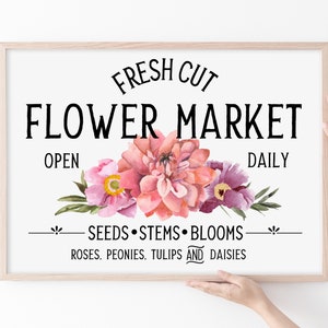 May include: A framed sign with the text "FRESH CUT FLOWER MARKET OPEN DAILY" and a floral design featuring pink, purple, and green flowers. The text below the flowers reads "SEEDS STEMS BLOOMS ROSES, PEONIES, TULIPS AND DAISIES."