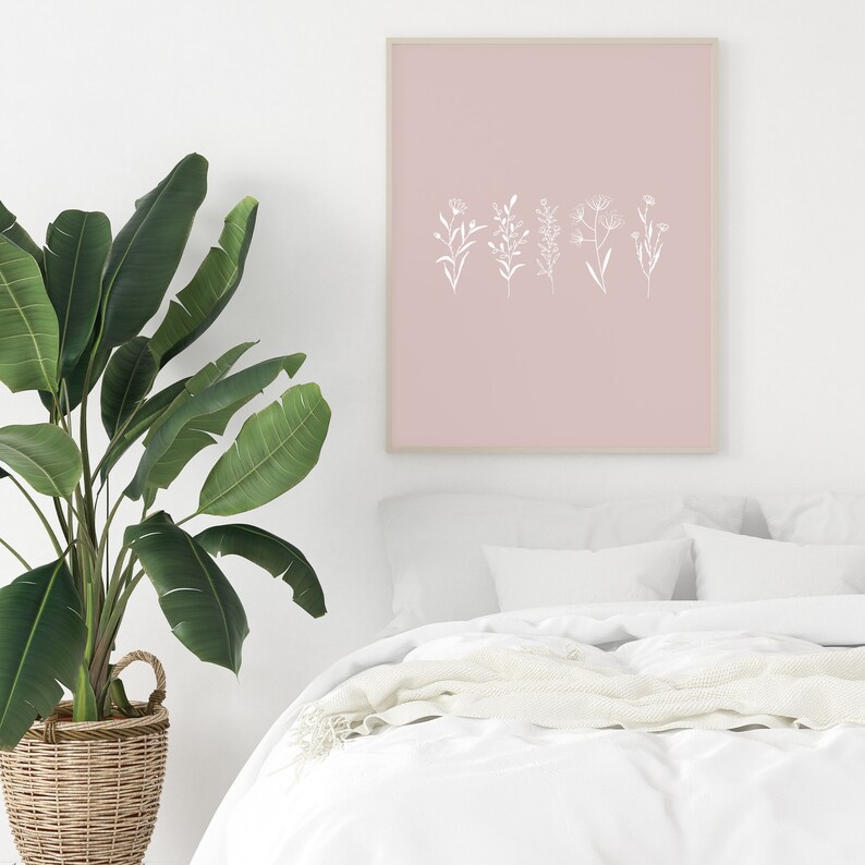 May include: A white framed print with a pink background and white line drawings of five different flowers. The print is hanging on a white wall above a white bed with a white blanket and pillows.