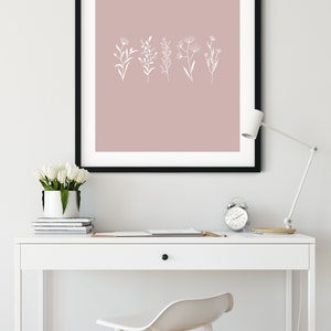 May include: A black framed print with a blush pink background and white line drawings of various flowers and plants. The print is hanging on a white wall above a white desk with a white chair and a white lamp.