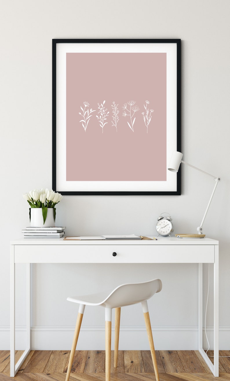 May include: A black framed print with a blush pink background and white line drawings of various flowers and plants. The print is hanging on a white wall above a white desk with a white chair and a white lamp.