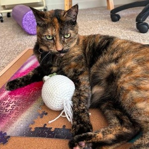 Puede incluir: Un gato carey con ojos verdes descansa sobre un rompecabezas de corcho. El gato es marrón, negro y naranja. Una bola blanca de ganchillo con hilos colgantes está cerca del gato. El rompecabezas tiene piezas moradas, rosas y negras.
