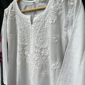Puede incluir: T&uacute;nica blanca de algod&oacute;n con intrincados bordados blancos con motivos florales y paisley. La t&uacute;nica tiene un escote en V y mangas tres cuartos.
