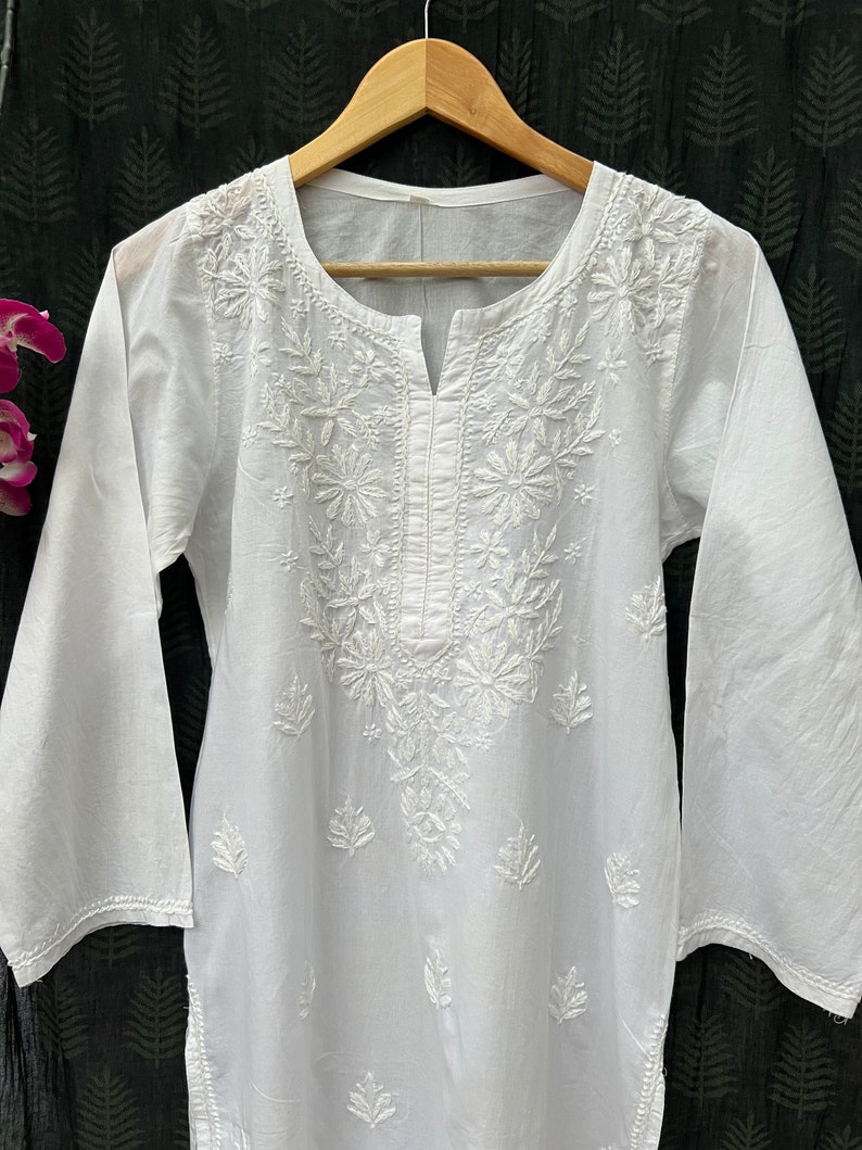 Puede incluir: T&uacute;nica blanca de algod&oacute;n con intrincados bordados blancos con motivos florales y de hojas. La t&uacute;nica tiene un escote redondo y mangas largas.