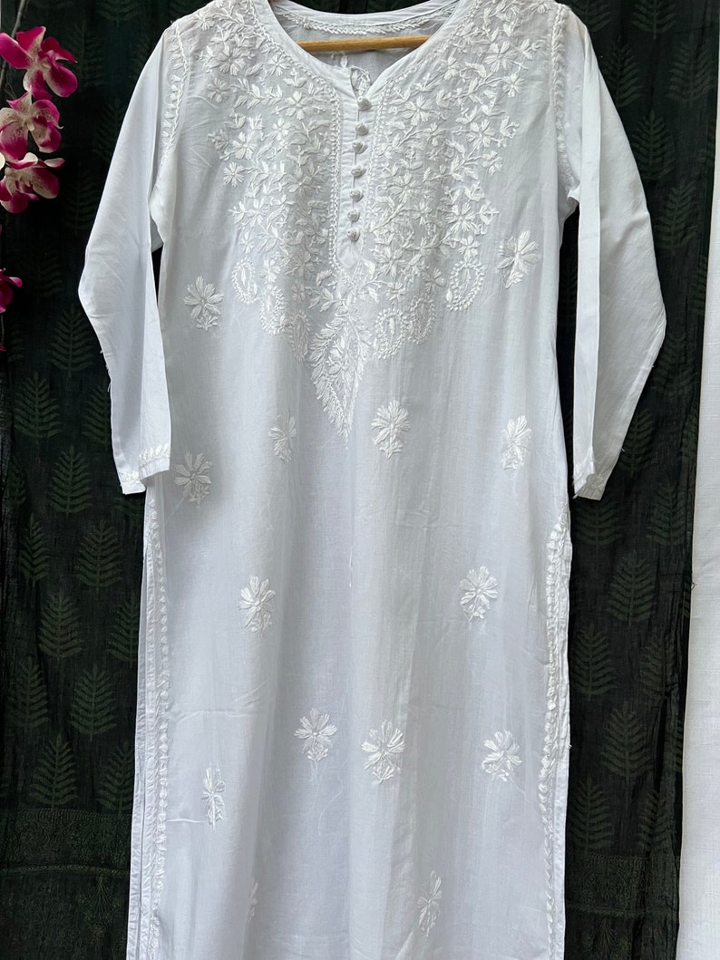 Puede incluir: T&uacute;nica blanca de algod&oacute;n con intrincados bordados blancos con dise&ntilde;os florales. La t&uacute;nica tiene un escote redondo y mangas largas.