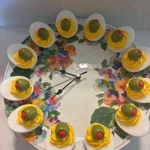 May include: A round wall clock featuring a floral pattern with pansies in shades of purple, pink, yellow, and green. Twelve deviled egg replicas are arranged around the perimeter, each topped with a green olive containing a red pimento.