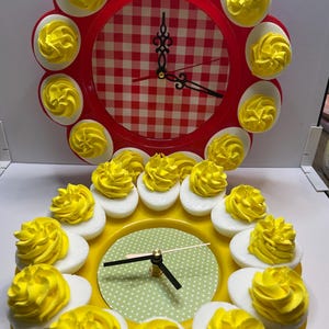 May include: Two decorative clocks with a food theme. The top clock has a red frame with a gingham pattern and is decorated with deviled egg halves. The bottom clock has a yellow frame and a green polka dot pattern, also decorated with deviled egg halves.
