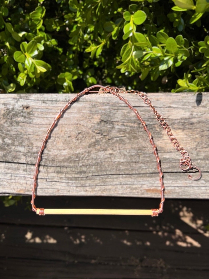 May include: A necklace featuring a horizontal, cylindrical, cream-coloured bead secured by copper wire wrapping. The necklace includes a copper wire chain and a metal extender chain with a circular clasp, showcasing a metallic copper finish throughout.