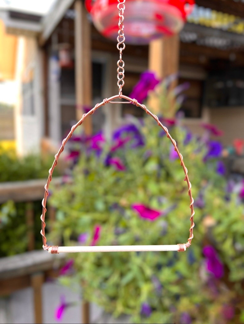 May include: A bird perch swing featuring a copper wire frame shaped into an arch with a twisted wire design. The swing includes a straight, light-coloured wooden dowel at the base, secured by coiled copper wire, and hangs from a copper chain.