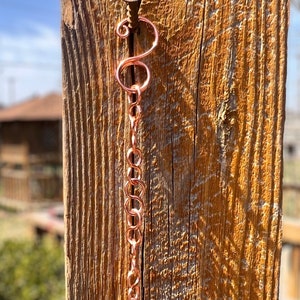 May include: A copper-coloured metal chain featuring a series of interlocking circular links, suspended from a single metal screw. The chain begins with a decorative S-shaped hook at the top, followed by a vertical arrangement of uniform round links.