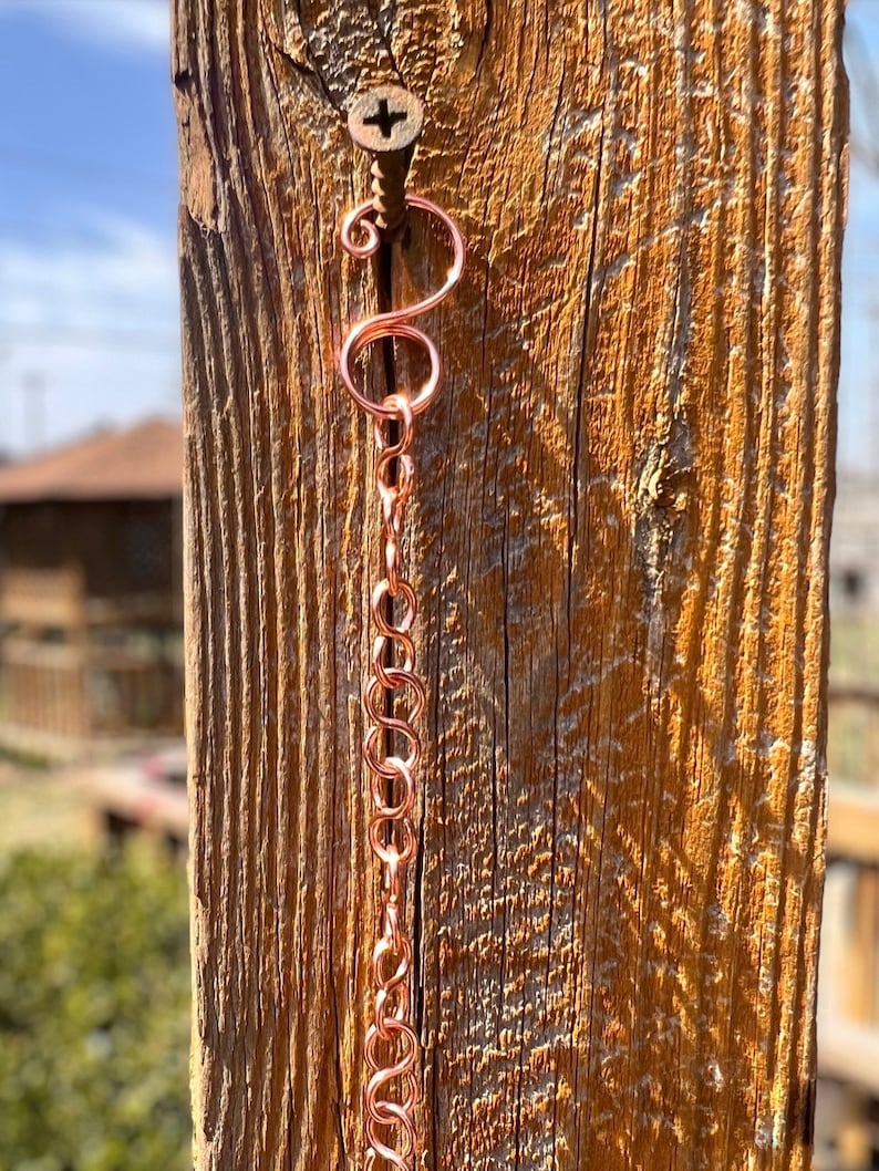 May include: A copper-coloured metal chain featuring a series of interlocking circular links, suspended from a single metal screw. The chain begins with a decorative S-shaped hook at the top, followed by a vertical arrangement of uniform round links.