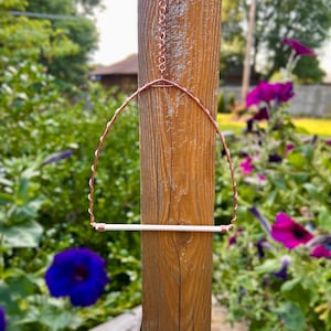 May include: A bird perch featuring a copper wire frame shaped into an arch with a twisted design. The base of the arch consists of a smooth, straight white dowel secured at both ends with copper wire coils. A copper chain is attached to the top.