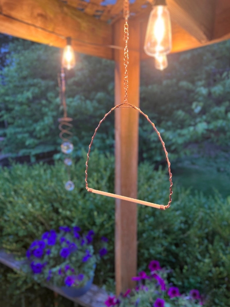 May include: A bird perch featuring a horizontal wooden dowel suspended by a twisted copper wire frame. The structure is attached to a metal chain for hanging, creating a triangular shape designed for avian use in an outdoor setting.