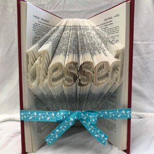 May include: A red hardback book with pages folded to spell out the word "Blessed". A light blue ribbon with a white leaf pattern is tied around the book.