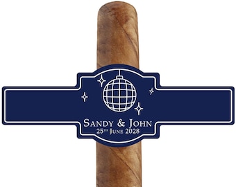 May include: A brown cigar with a navy blue label. The label features a disco ball graphic and the text "SANDY & JOHN 25th JUNE 2028". The cigar is set against a white background.