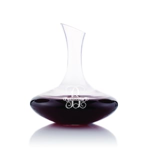 May include: A clear glass wine decanter with a wide base and a narrow neck. The decanter is filled with red wine and has the monogram "JB" with the text "BECKHAM" engraved on the side.
