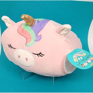 Peut inclure: Une peluche licorne rose avec une corne dorée et des détails de crinière colorés. La licorne a les yeux fermés et une étiquette attachée. La peluche est conçue pour être douce et câline.