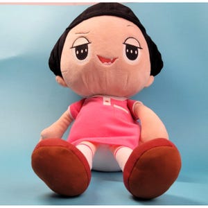 May include: A plush doll with a smiling face, black hair, and large eyes. The doll is wearing a pink dress with white trim and brown shoes. The doll is sitting with its legs outstretched.