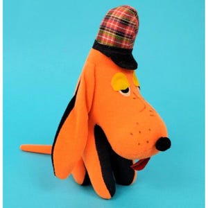 Könnte beinhalten: Orangefarbenes Plüschhundespielzeug mit schwarzen Akzenten, einer roten Zunge und einer karierten Mütze. Der Hund hat gelbe Augen und eine lange Schnauze. Das Spielzeug befindet sich vor einem einfarbigen blauen Hintergrund.