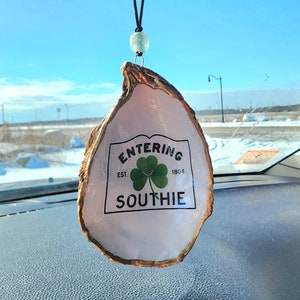 May include: A white oyster shell car charm with a gold trim. The shell has a sign that reads "Entering Est. 1804 Southie" with a green shamrock.