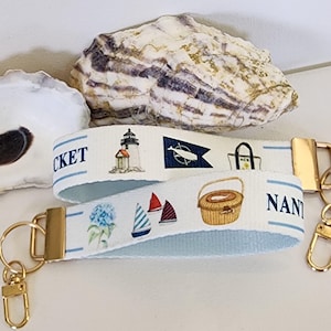 May include: A white fabric keychain wristlet featuring a repeating nautical print of a lighthouse, a whale flag, a tote bag, sailboats, a basket, and blue hydrangeas. The text NANTUCKET is printed in blue, finished with gold metal hardware.