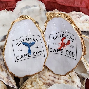 May include: Two white oyster shells with gold-painted edges. Each shell features a black and white label with the text "ENTERING CAPE COD" and the year "1602". One label has a blue whale tail, the other a red lobster.
