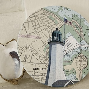 May include: A round coaster featuring a lighthouse illustration with the word "Scituate" and a map. The lighthouse is white with a black top and a small American flag. The coaster is placed next to a white oyster shell.
