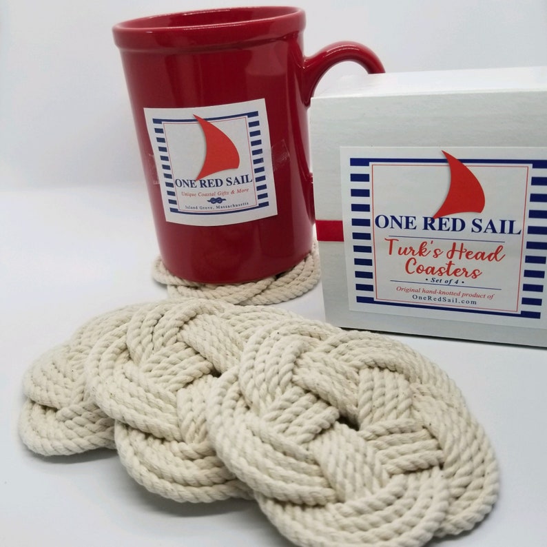 K&ouml;nnte beinhalten: Set aus vier wei&szlig;en Seiluntersetzern mit einem T&uuml;rkischen Kopf-Knotenmuster. Die Untersetzer sind in einer wei&szlig;en Box mit einem blau-wei&szlig; gestreiften Etikett verpackt, auf dem "ONE RED SAIL Turk's Head Coasters - Set of 4 - Original hand-knotted product of OneRedSail.com" steht.