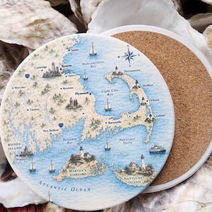 May include: Round ceramic coaster featuring a detailed map of Cape Cod, Massachusetts, and surrounding areas. The map includes nautical elements like sailboats, lighthouses, and a compass rose, with place names in a blue and beige color scheme. The coaster has a cork backing.