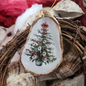 May include: A white oyster shell with a gold rim, decorated with a painted Christmas tree with red bows and white ornaments. The ornament is hanging from a gold string.
