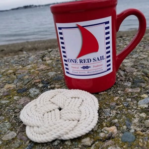 K&ouml;nnte beinhalten: Eine rote Keramiktasse mit einem wei&szlig;en Logo, auf dem "ONE RED SAIL" steht, darunter die Worte "Nautical Traditions" und "Island Grove, Massachusetts". Die Tasse steht auf einem wei&szlig;en Untersetzer aus Seil.