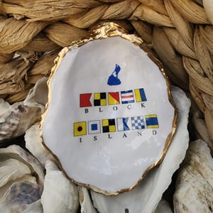 May include: A white oyster shell dish with a gold rim, featuring the words "BLOCK ISLAND" in colorful nautical signal flags. A blue map of Block Island is also present.