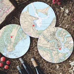 May include: Three round coasters featuring nautical charts of coastal areas, each with a red heart marking a location. The coasters are arranged on a bed of straw, with wine bottles and red berries in the foreground. The charts are in shades of blue, beige, and green.