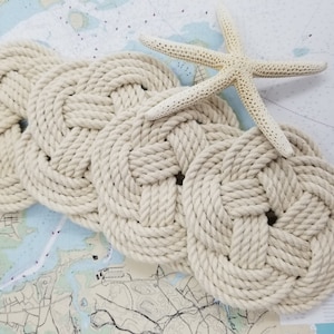 K&ouml;nnte beinhalten: Ein Set aus drei geflochtenen Korkuntersetzern in nat&uuml;rlicher Cremefarbe. Die Untersetzer sind in Wellenform angeordnet und aus dickem, geflochtenem Seil gefertigt.