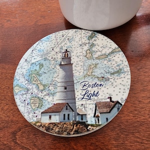 May include: Round coaster featuring a detailed illustration of the Boston Light lighthouse, set against a nautical chart background. The coaster has the words "Boston Light" in a script font. The coaster is on a wooden surface.