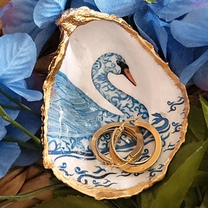 May include: A decorative oyster shell trinket dish featuring a hand-painted blue and white swan design on the interior. The edges of the shell are finished with a metallic gold trim. Two small gold hoop earrings rest inside the shell dish.
