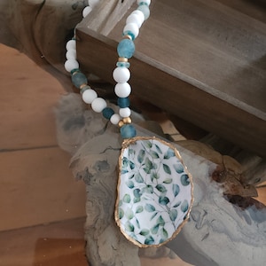 May include: A white and blue beaded necklace with a gold-rimmed oyster shell pendant featuring a green leafy design.